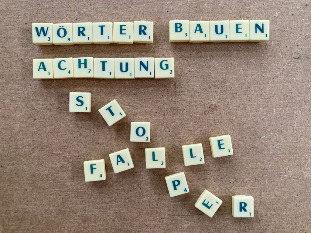 Text mit Buchstabensteinen von Scrabble gelegt, er heisst: Wörter bauen Achtung Stolperfalle. Beim Wort Stolperfalle geraten die Buchstaben durcheinander