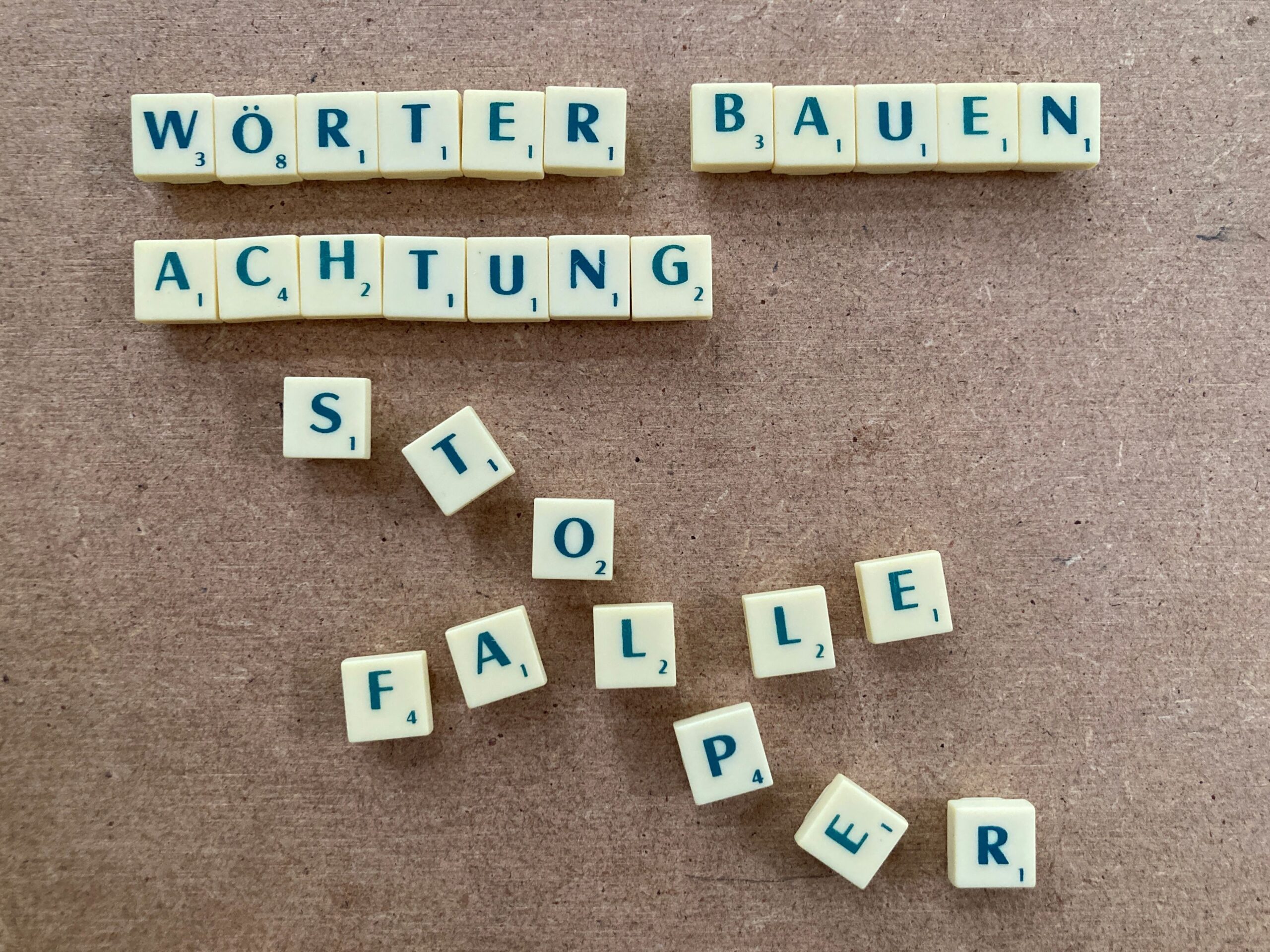 Text mit Buchstabensteinen von Scrabble gelegt, er heisst: Wörter bauen Achtung Stolperfalle. Beim Wort Stolperfalle geraten die Buchstaben durcheinander